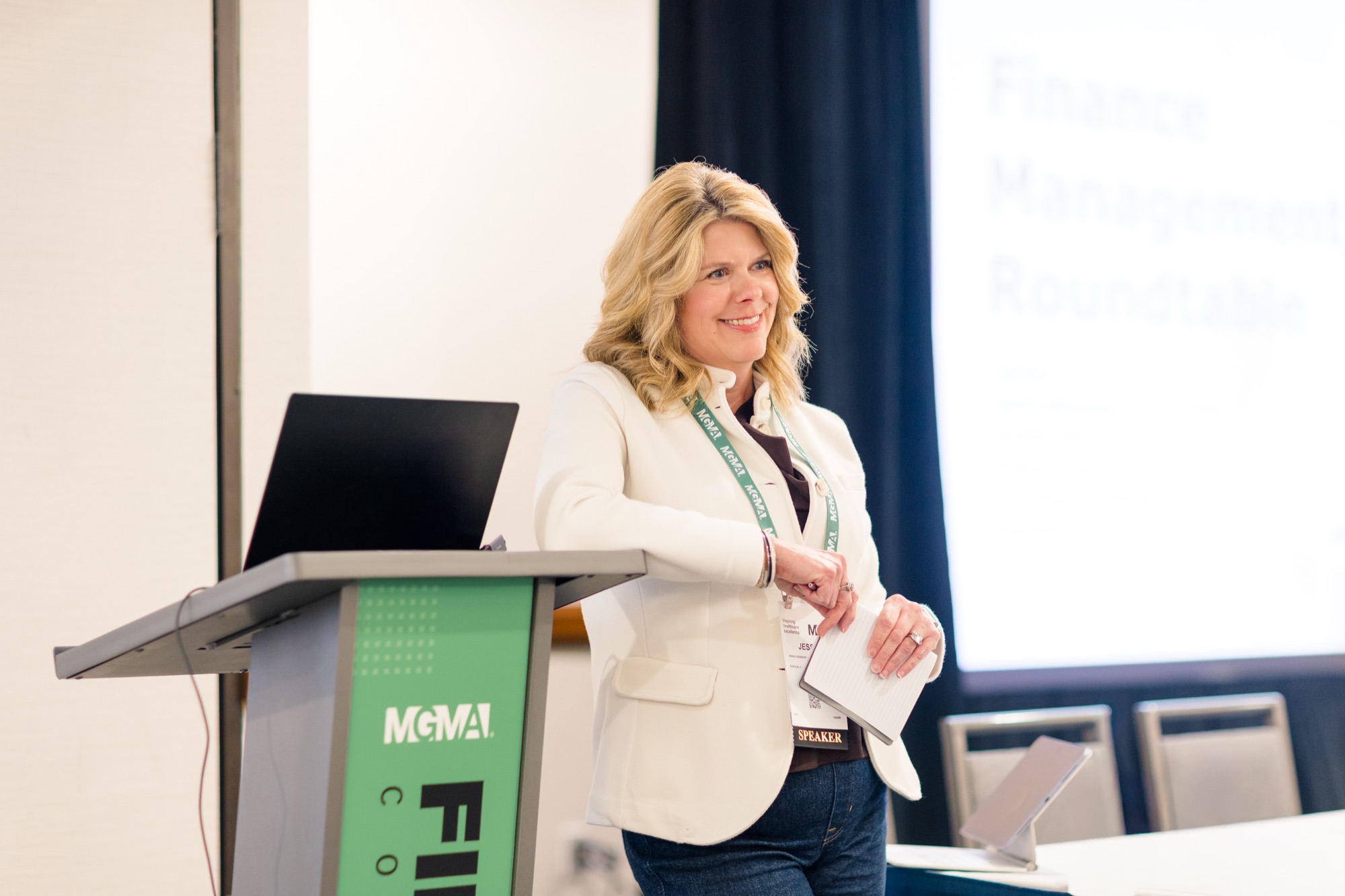 Jessica Minesinger smiling beside a podium while speaking at an MGMA conference